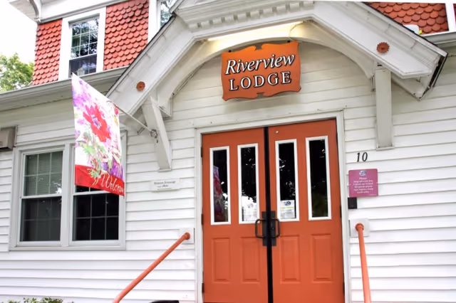 Front entrance of Riverview Lodge Residential Care Home featuring double orange doors with glass panels, a wooden sign above the door with the name 'Riverview LODGE', white siding, a window to the left, a colorful floral welcome flag, and a small purple sign on the right side of the door.