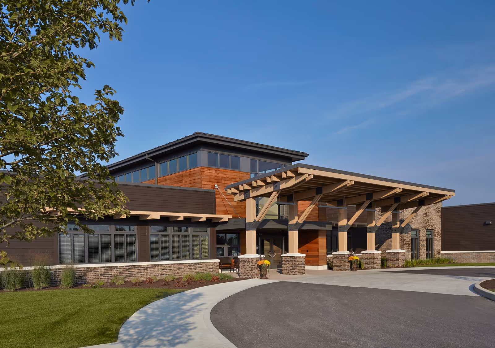Exterior view of a modern senior living facility building with a covered entrance, large windows, and a mix of brick and wood siding under a clear blue sky.