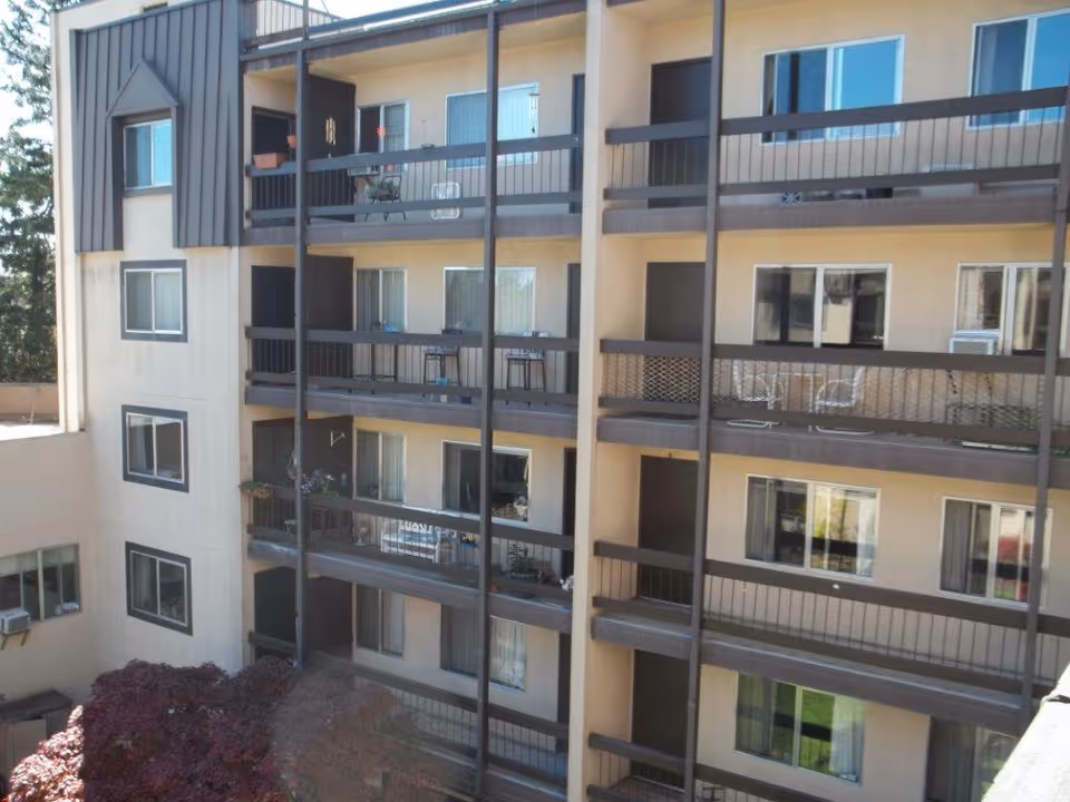 Exterior view of a multi-story retirement center building with balconies on each floor. The balconies have railings and some have chairs, tables, and potted plants. Windows and sliding glass doors are visible on each floor. There are trees and bushes near the base of the building.