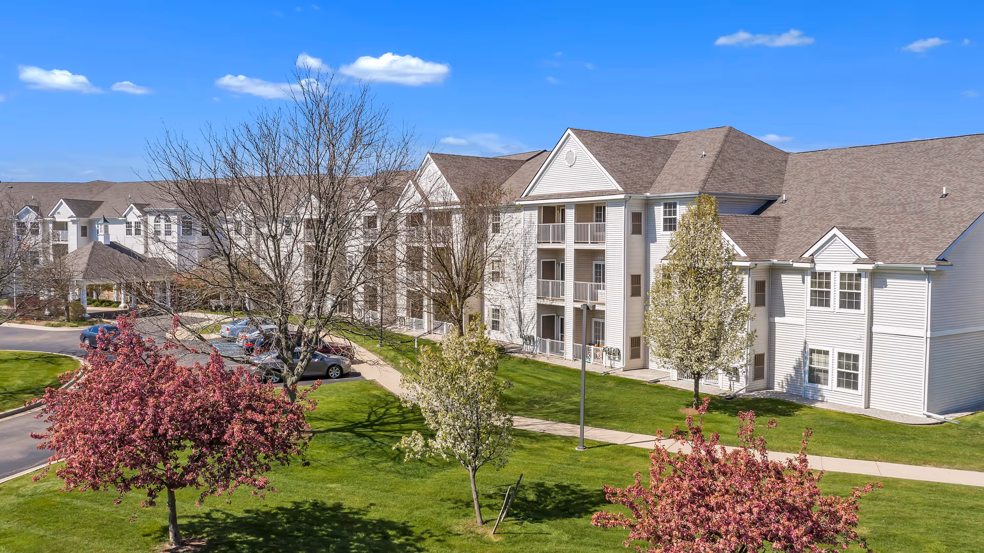Exterior view of a large, multi-story senior living facility building with white siding and multiple balconies. The building is surrounded by green lawns, blossoming trees with pink and white flowers, and a parking area with several cars. The sky is clear and blue with a few clouds.