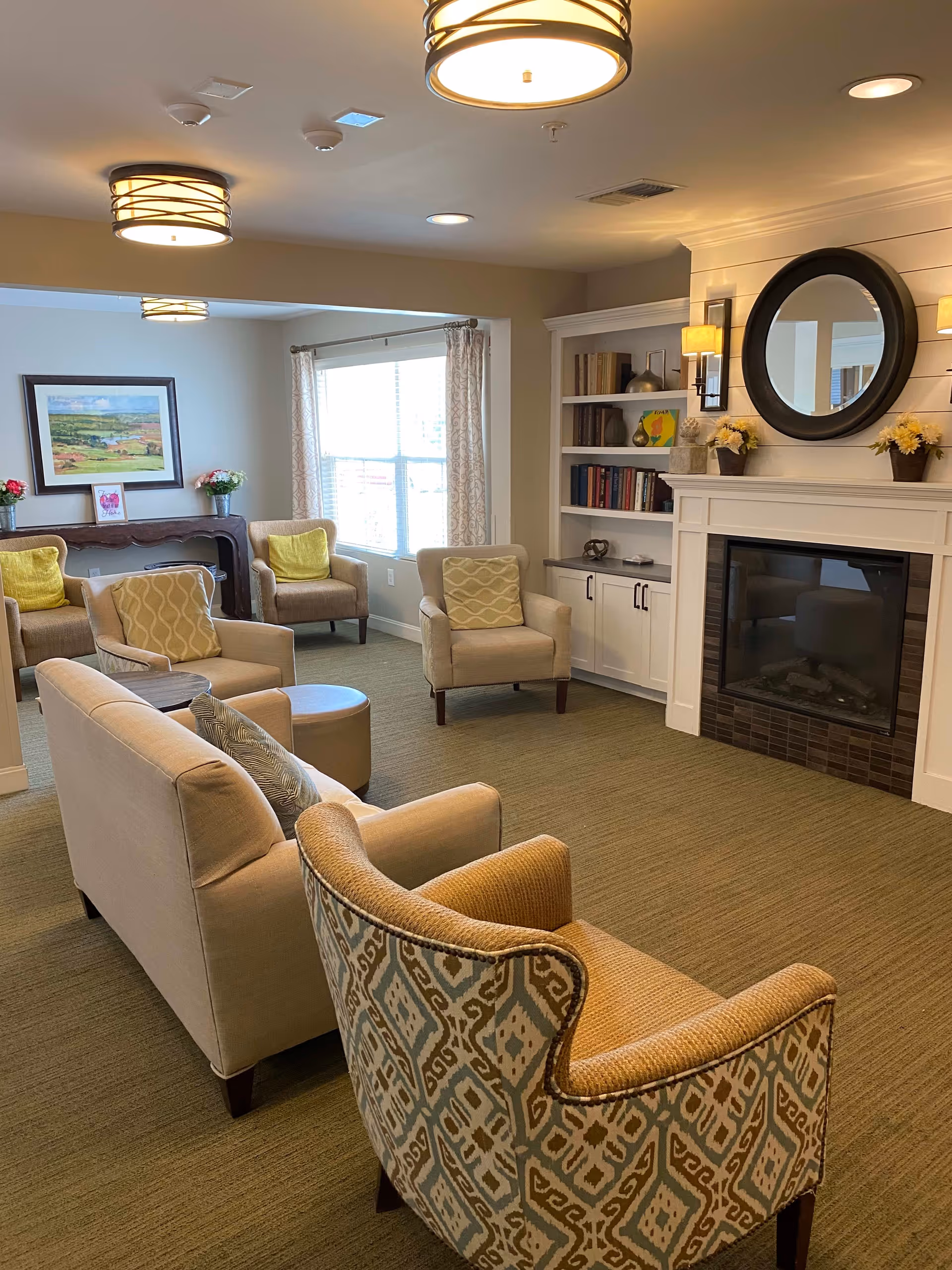 A cozy living room area with beige and patterned armchairs, a beige sofa, a round ottoman, and a fireplace with a round mirror above it. Built-in white shelves with books and decorative items flank the fireplace. There is a large window with curtains letting in natural light, and a framed landscape painting on the wall. The room has warm lighting from ceiling fixtures and wall sconces.