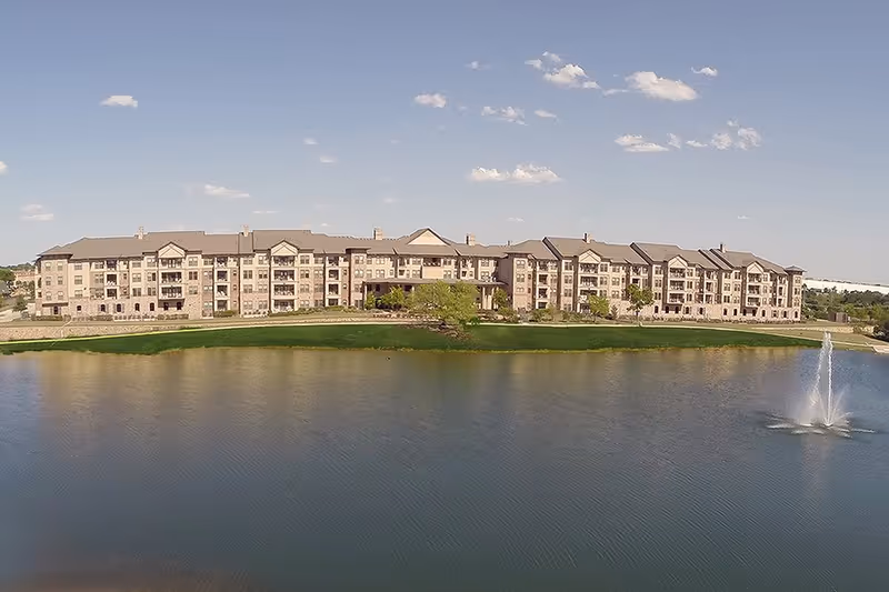 A large multi-story senior living facility building situated behind a pond with a water fountain, under a partly cloudy blue sky.