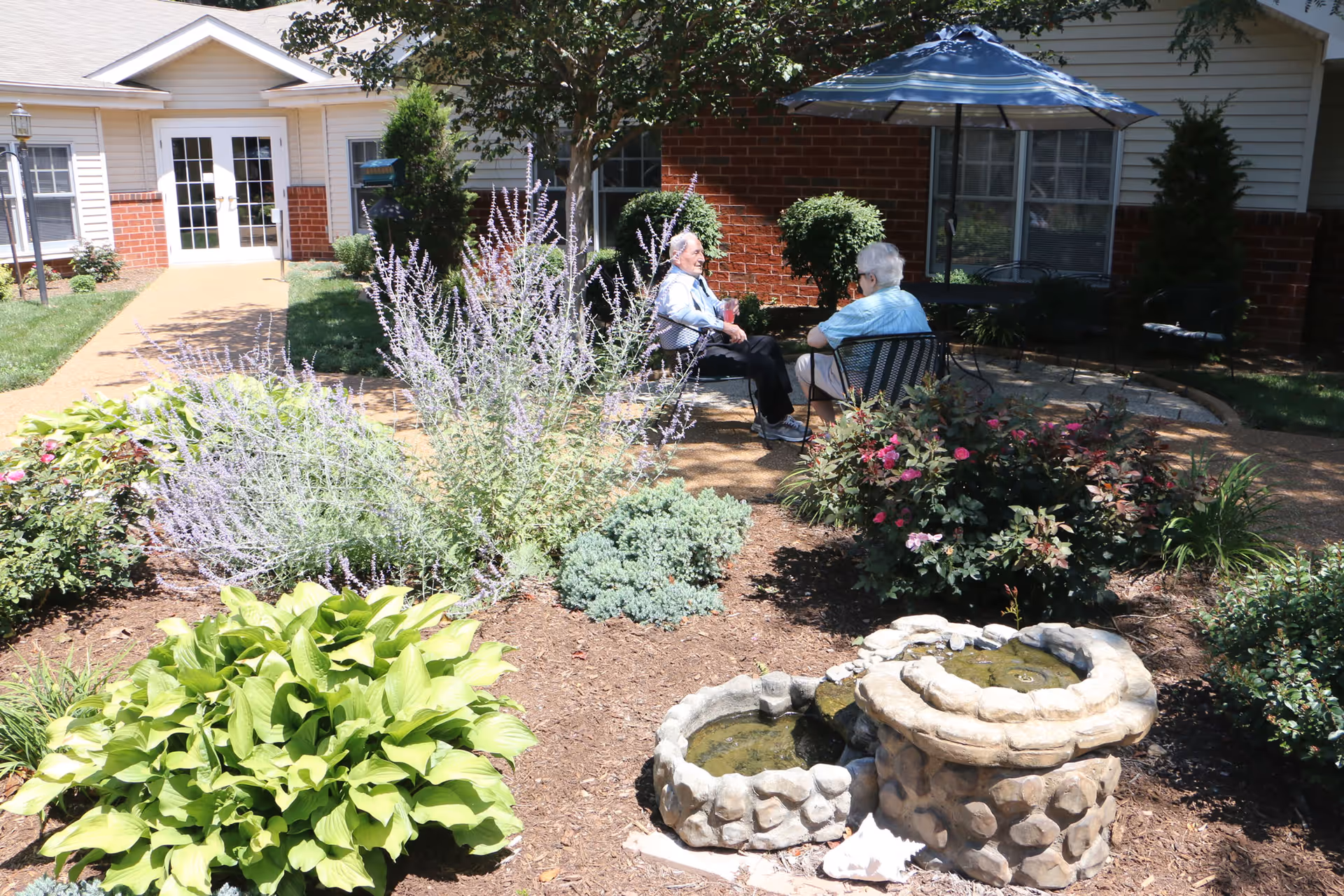 Two elderly people sitting and talking on metal chairs in a garden patio area with flowering plants, shrubs, and a small stone water fountain. The background shows a brick and siding building with a door and windows, and a blue umbrella providing shade.