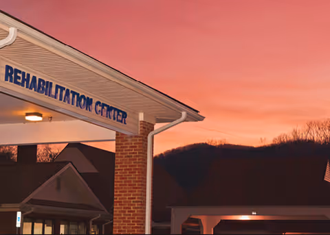 Exterior view of a rehabilitation center building at sunset with a pink and orange sky and silhouetted hills in the background. The building has a brick column and a covered entrance with the words 'REHABILITATION CENTER' visible.