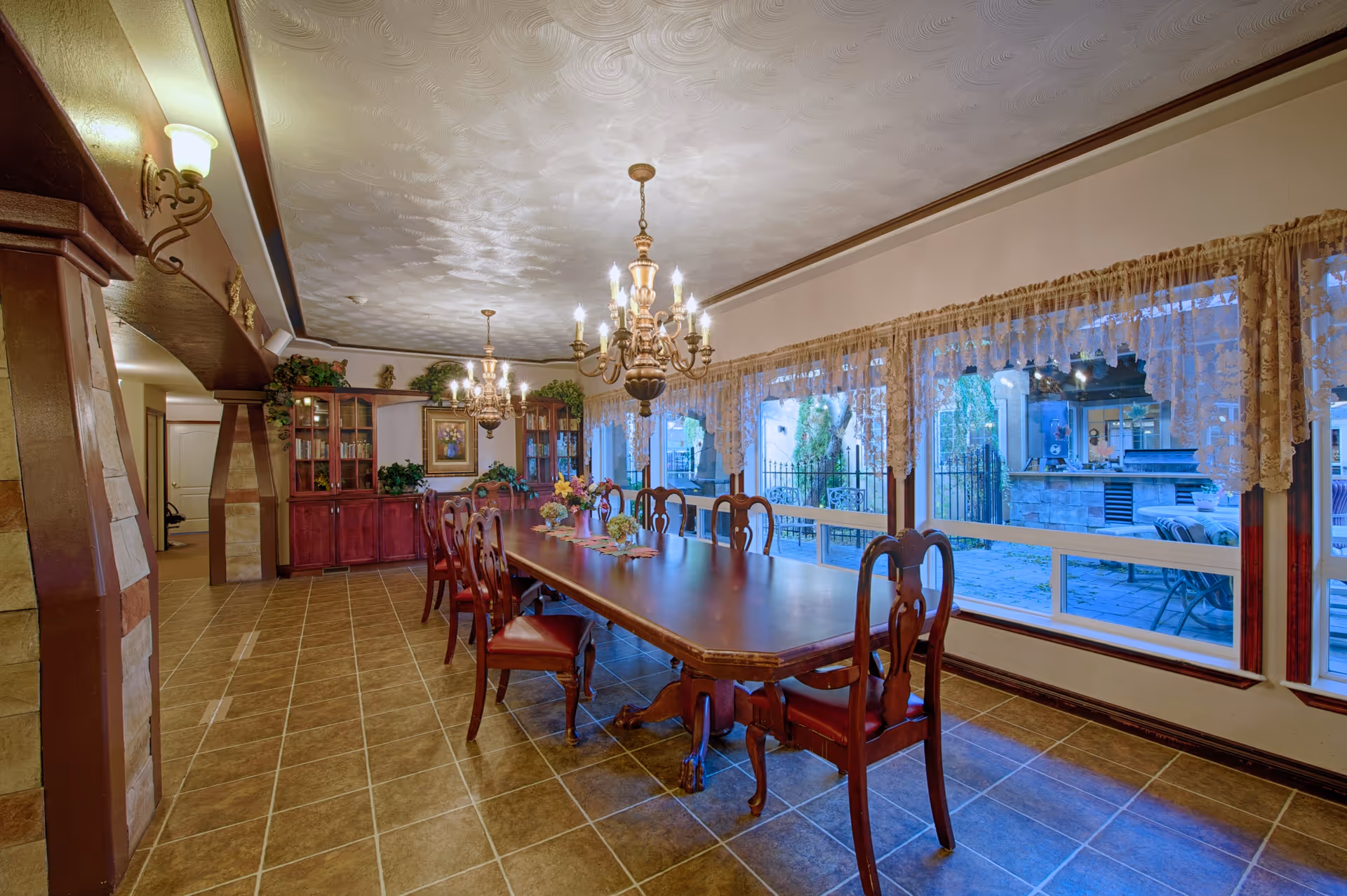 A spacious dining room with a long wooden table surrounded by wooden chairs with red cushions. Two ornate chandeliers hang from a textured ceiling. Large windows with lace curtains line one side of the room, offering a view of an outdoor patio area. A wooden cabinet with glass doors and decorative plants is against the far wall.