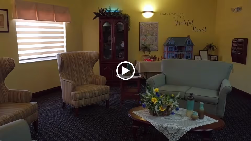 A cozy living room area with two striped armchairs, a light blue loveseat, a wooden coffee table with a lace doily and a flower arrangement, a wooden cabinet with glass doors, and a table with decorative items including a dollhouse. The walls are painted yellow with a window covered by a beige blind and a wall decal that reads 'Begin each day with a grateful heart.'