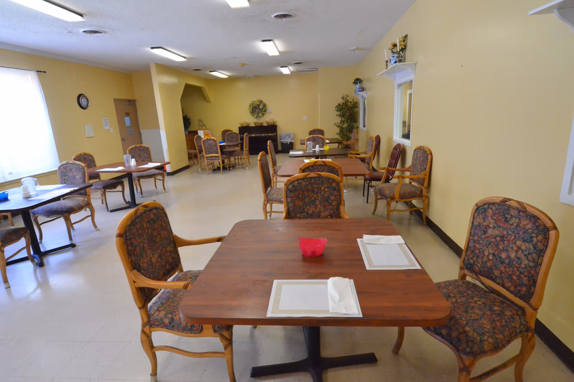 A dining room in Hermitage Retirement Center with several wooden tables and floral upholstered chairs arranged neatly. Each table has placemats and napkins, with some tables having pitchers and small bowls. The walls are painted yellow, and there is a clock on one wall and some decorative plants and wall hangings in the background.