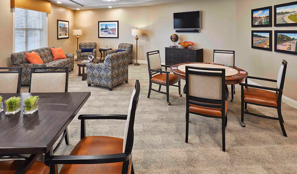 A well-lit common room with patterned armchairs and a sofa with orange cushions arranged around a coffee table, a round table with chairs and poker chips, a flat-screen TV mounted on the wall above a black dresser, and framed pictures on the walls.