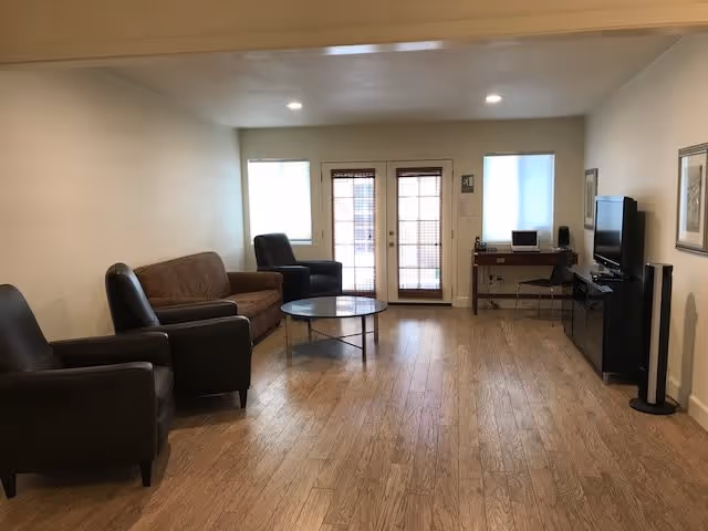 A living room with wooden flooring featuring a brown sofa, two black armchairs, a round glass coffee table, a desk with a chair and computer, a flat-screen TV on a black TV stand, and two windows with blinds flanking a glass door.