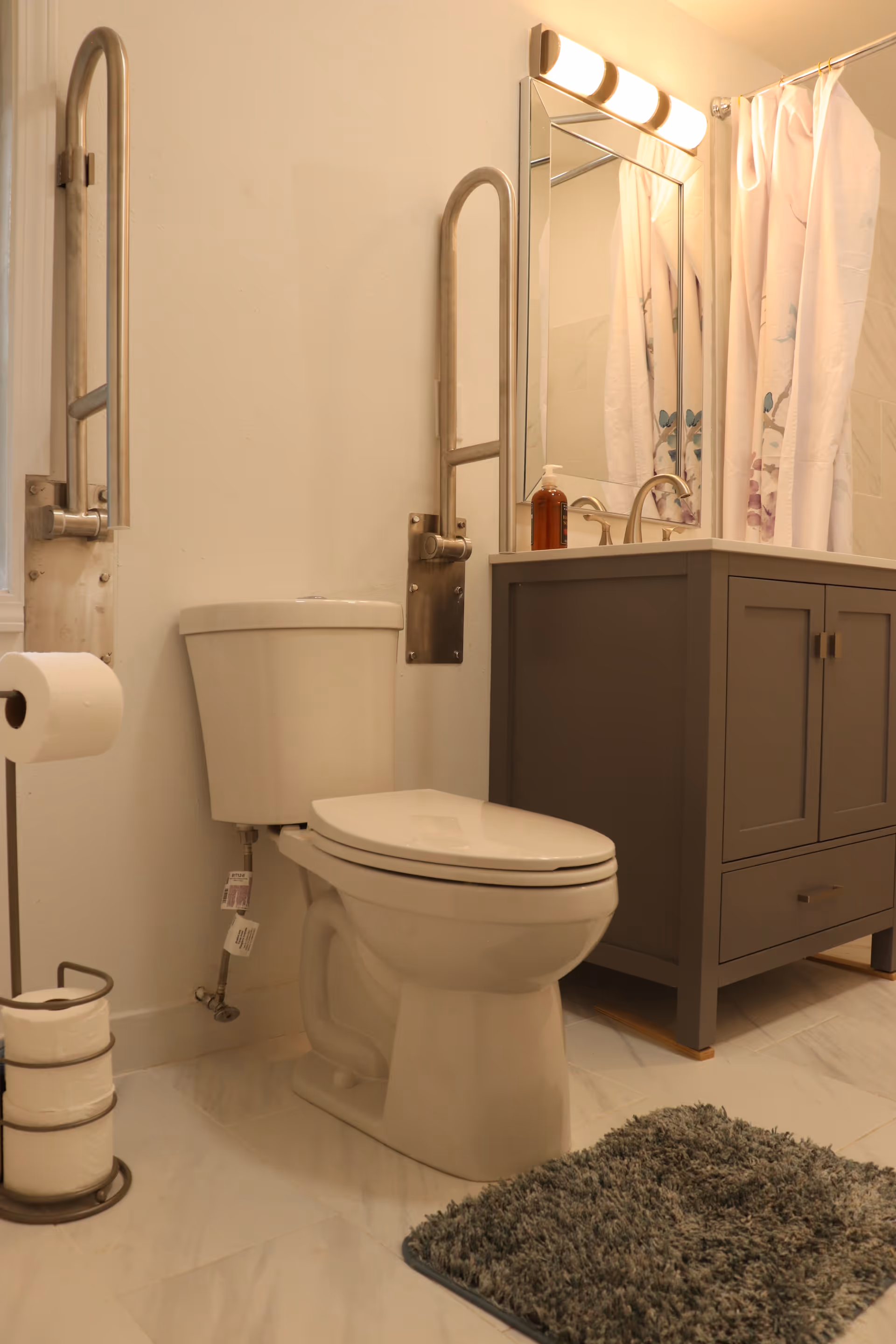 A clean bathroom featuring a white toilet with safety grab bars on both sides, a gray vanity with a sink and mirror above it, a bottle of soap on the vanity, a shower curtain with floral patterns, a toilet paper holder with extra rolls, and a gray bath mat on the tiled floor.