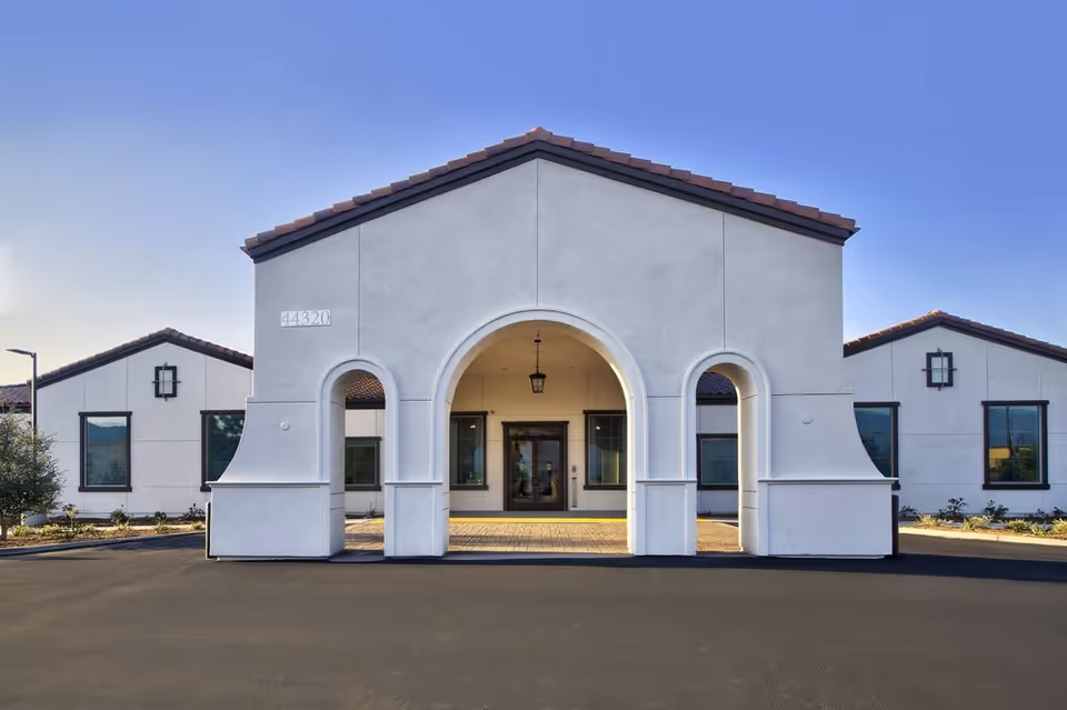 Front exterior view of Temecula Memory Care facility featuring a white building with a tiled roof and three large archways at the entrance, surrounded by a paved driveway and landscaped areas with small plants and trees under a clear blue sky.
