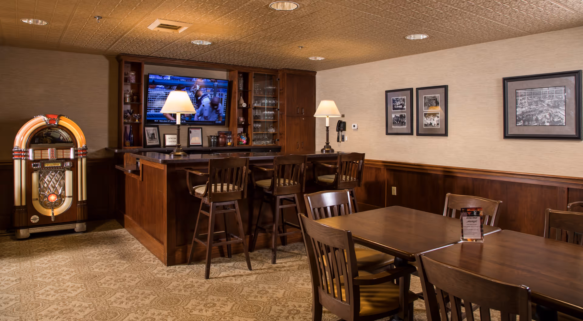 A cozy interior room with a wooden bar counter and four bar stools. A vintage jukebox stands to the left of the bar. Behind the bar is a mounted flat-screen TV showing a program with a dog. The room has warm lighting with two table lamps on the bar counter. There are framed black and white photos on the beige wall, and a wooden dining table with chairs is in the foreground.