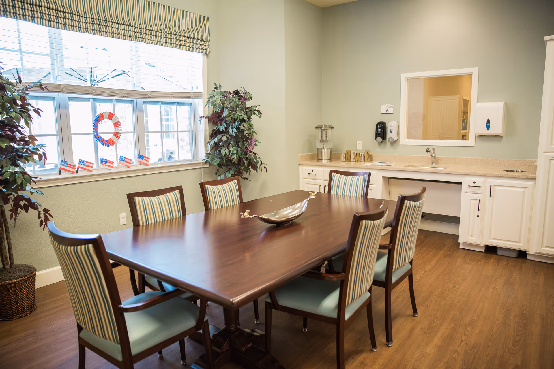 A dining room with a rectangular wooden table surrounded by six chairs with striped upholstery. There are two potted plants near a large window decorated with small American flags and a red, white, and blue wreath. The room has light green walls, a wooden floor, and a countertop with cabinets, a sink, and dispensers on the right side.