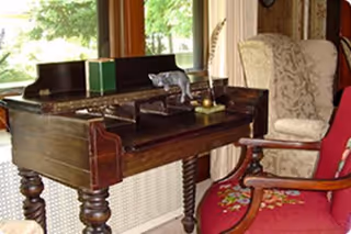 A cozy interior corner featuring a dark wooden antique desk with turned legs, placed near a window with green trees visible outside. On the desk are a few small decorative items. Next to the desk is a cushioned armchair with a floral pattern and a red upholstered chair with wooden armrests.