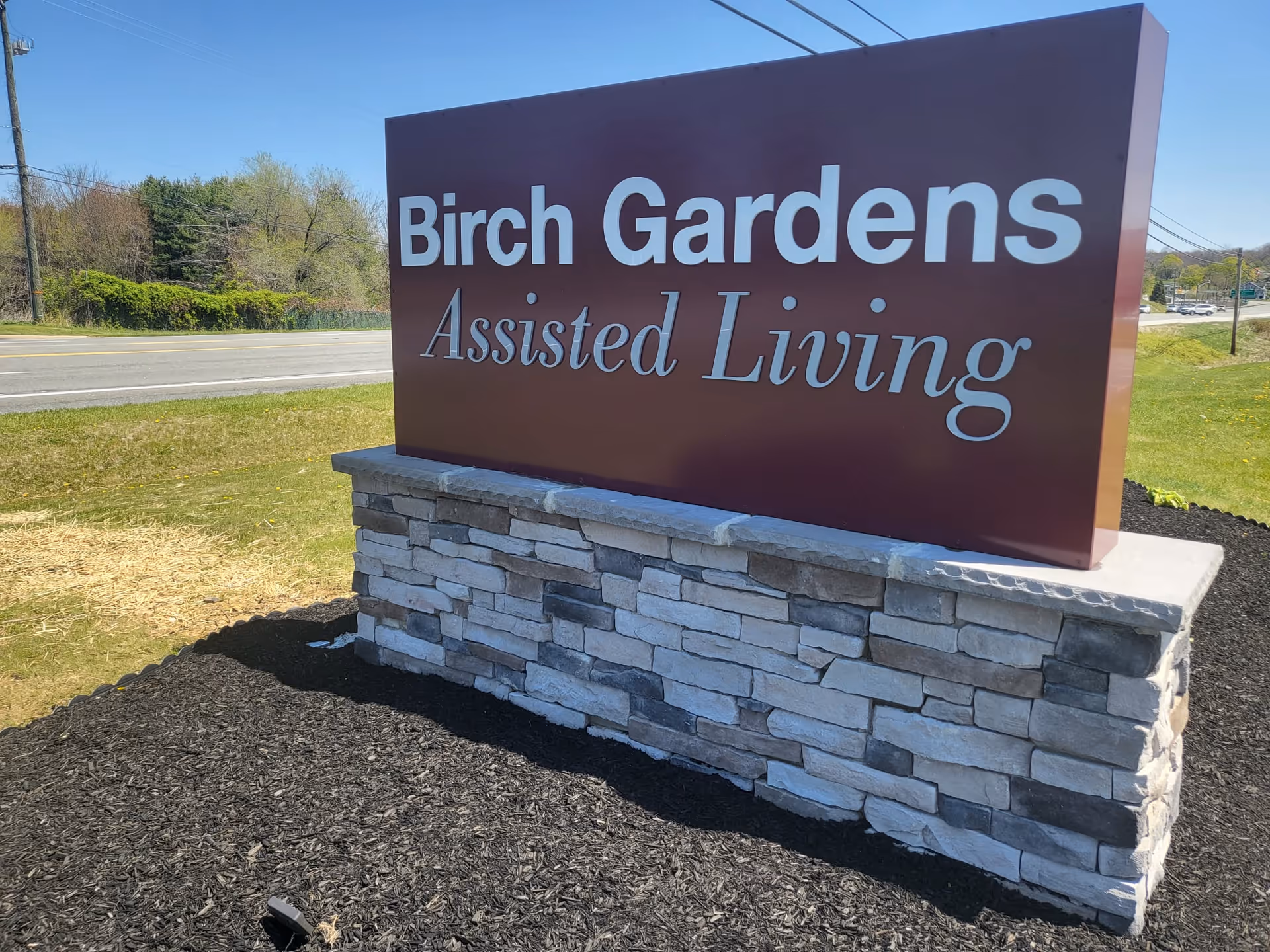 Large outdoor sign reading 'Birch Gardens Assisted Living' mounted on a stone base beside a road.