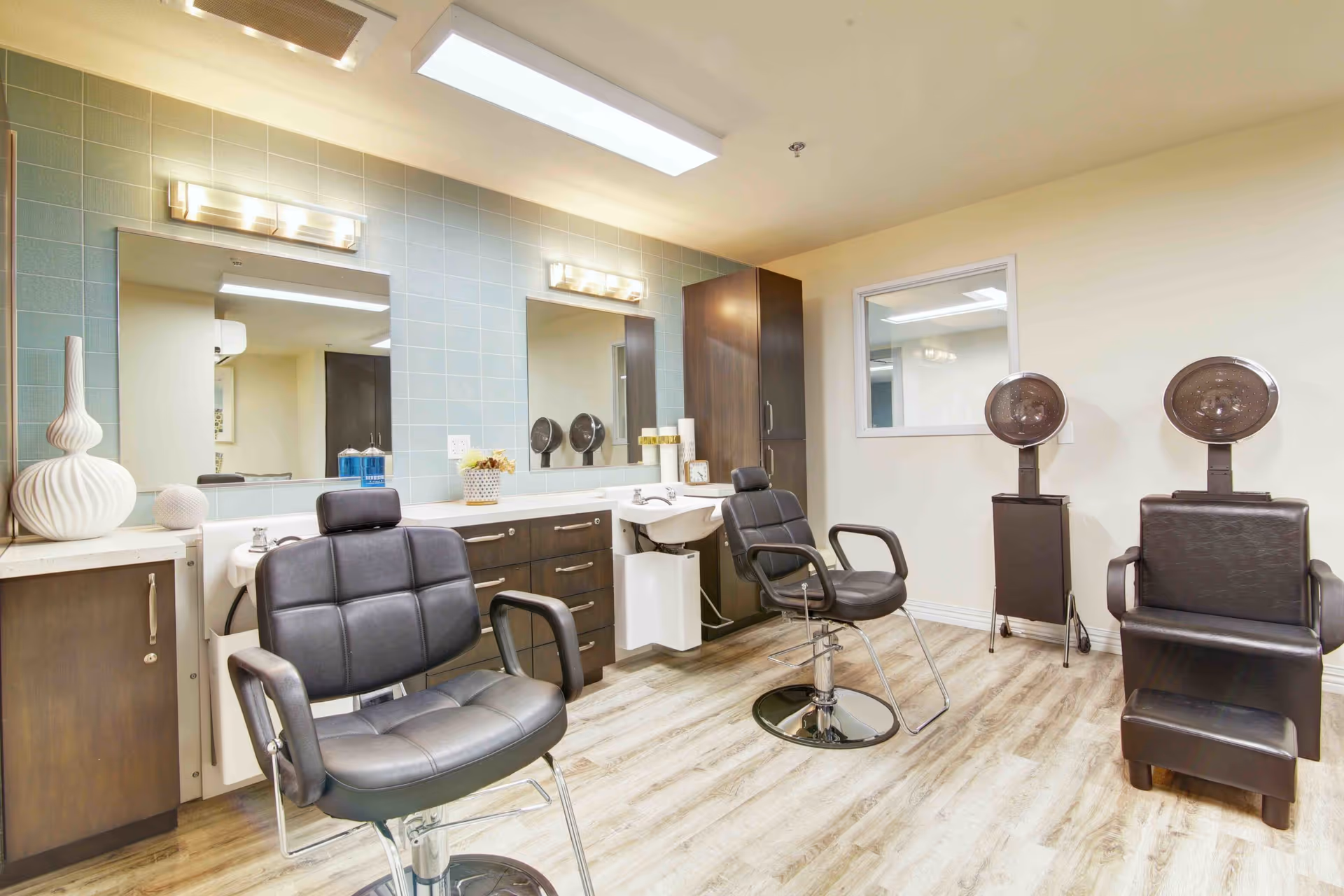 Interior view of a salon area in a senior living facility featuring two black salon chairs in front of a counter with sinks and mirrors, and two hair dryers on stands against a beige wall. The floor has a light wood finish and the walls are light-colored with a tiled backsplash behind the counter.