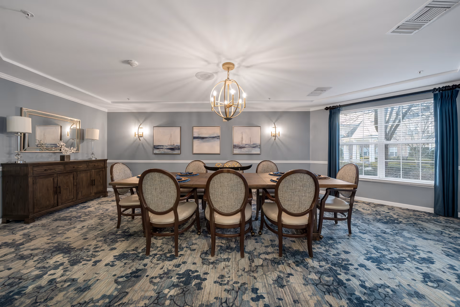 A spacious dining room with a large wooden table surrounded by eight upholstered chairs. The room features a patterned carpet, a modern chandelier hanging from the ceiling, three framed paintings on the wall, a large mirror above a wooden sideboard, and a large window with blue curtains letting in natural light.