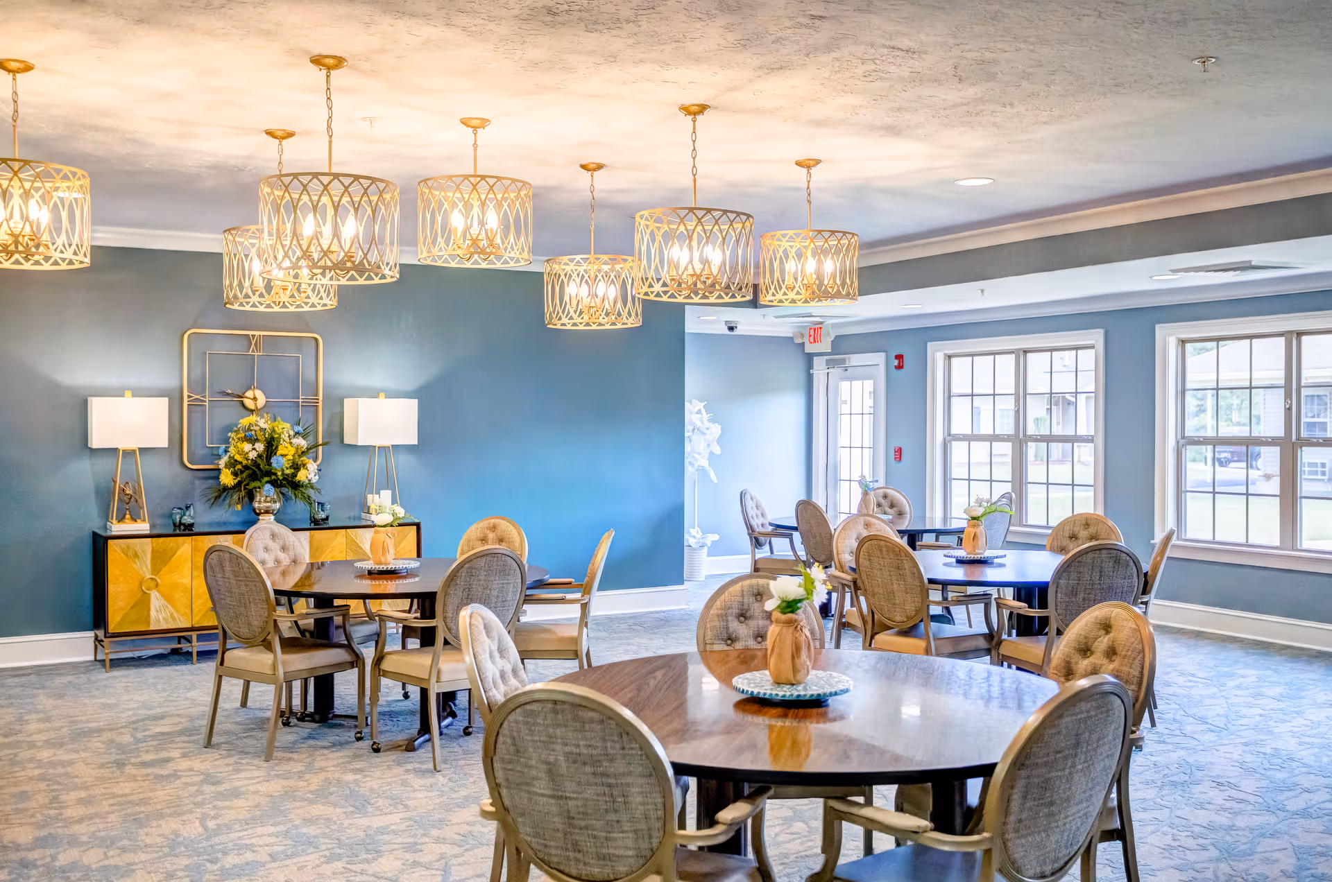 A bright and spacious dining room with multiple round wooden tables surrounded by cushioned chairs. The room features large windows allowing natural light to fill the space, blue walls, and decorative hanging light fixtures. A sideboard with lamps and a floral arrangement is against one wall.