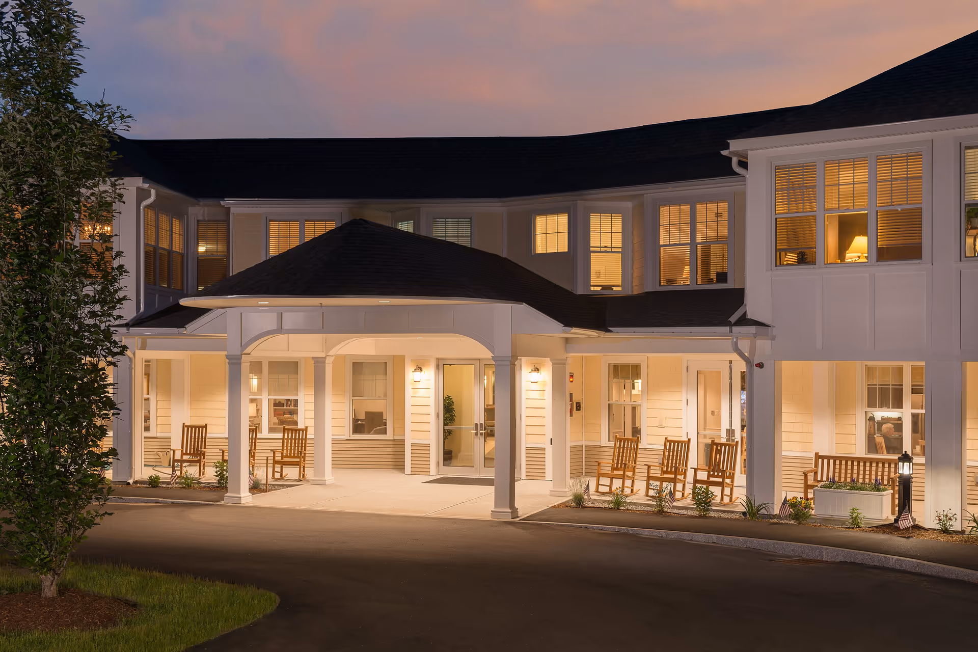 Exterior view of the Scott-Farrar at Peterborough senior living facility at dusk, showing a well-lit entrance with a covered porch, several wooden rocking chairs, and windows revealing warmly lit interior rooms.