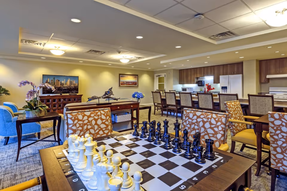 A spacious common area in a senior living facility featuring a large chessboard with pieces set up on a table in the foreground. The room includes comfortable seating with patterned chairs, a television on a wooden cabinet, decorative plants, and a kitchen area with a long counter and multiple chairs in the background. The lighting is warm and the ceiling has recessed lights and panels.