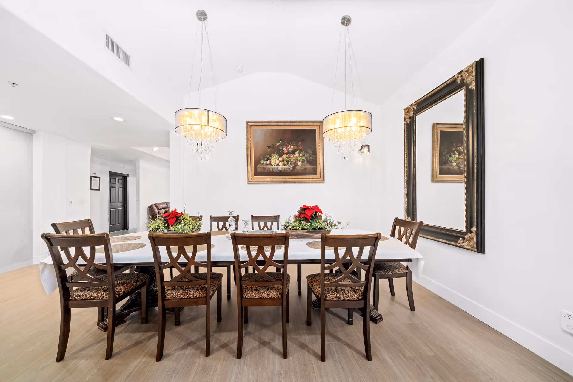 A bright dining room with a long rectangular table covered with a white tablecloth and surrounded by eight wooden chairs with patterned cushions. Two elegant chandeliers hang above the table. On the table are two floral centerpieces with red flowers. The room has light-colored wooden flooring, white walls, a large ornate mirror on the right wall, and a framed painting of fruit on the back wall.