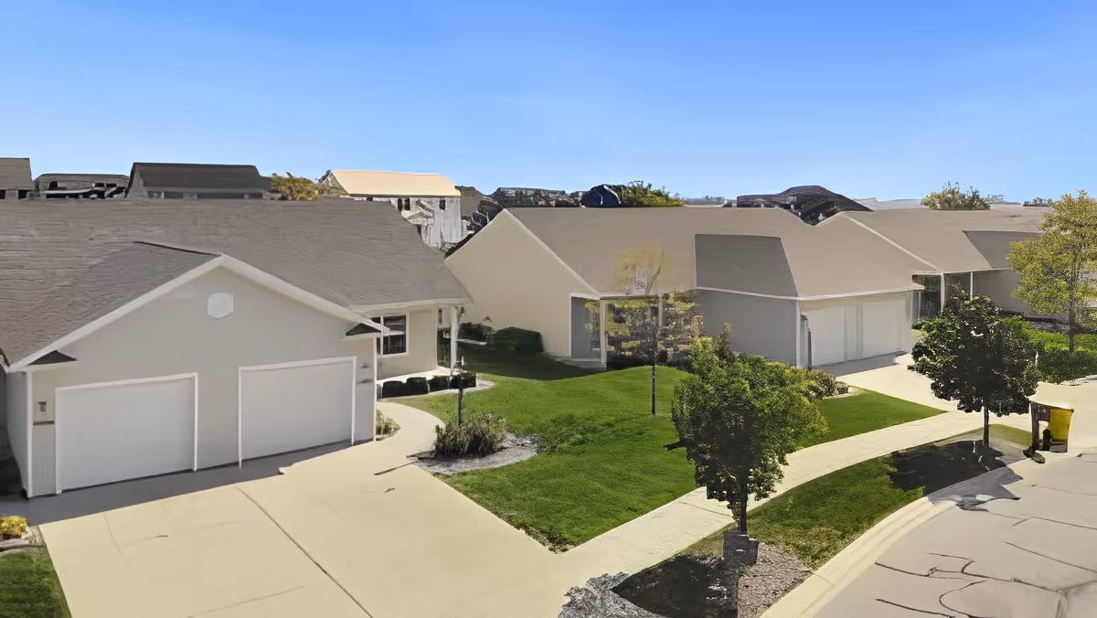 Exterior view of a senior living facility showing multiple single-story buildings with garages, green lawns, trees, and paved walkways under a clear blue sky.