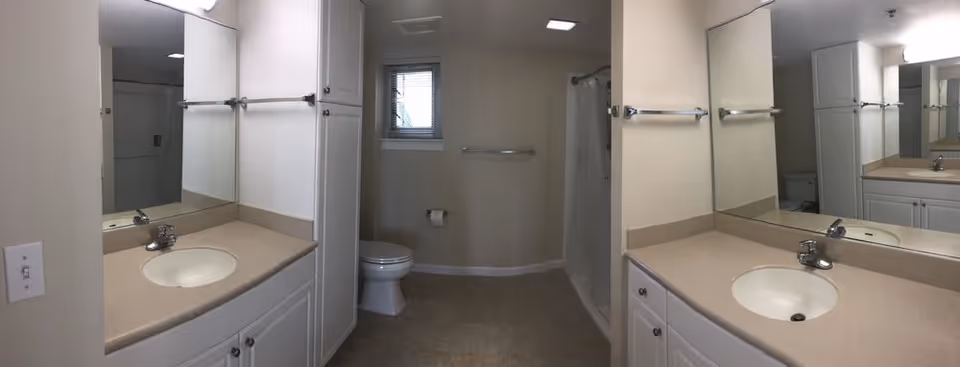 A clean, well-lit bathroom featuring a beige countertop with two sinks and chrome faucets, white cabinetry, a toilet, a small window with blinds, a towel rack, and a shower area with a curtain.