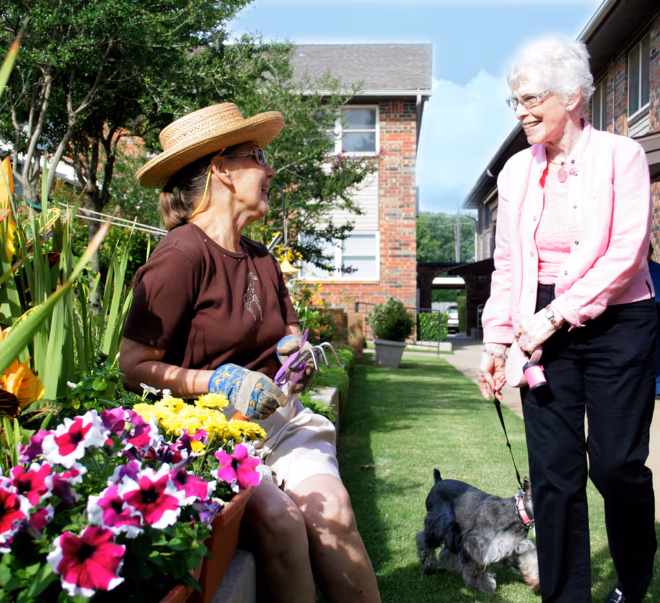 Two elderly women enjoying time outdoors in a garden area of a retirement community. One woman wearing a straw hat and gardening gloves is sitting and holding gardening tools near colorful flowers, while the other woman, dressed in a pink jacket, is standing and smiling while holding a small dog on a leash. They are surrounded by greenery and brick buildings.