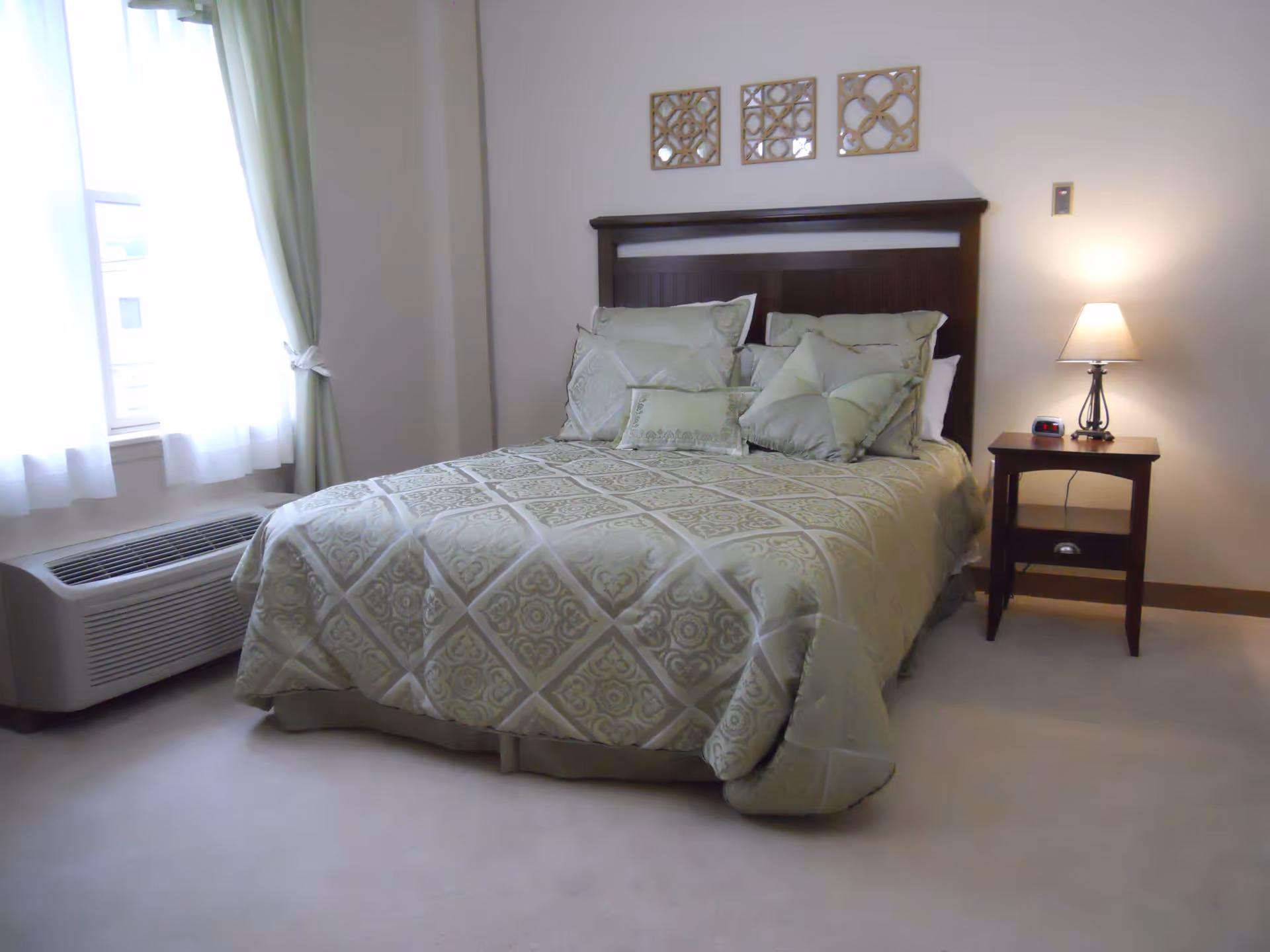 A neatly made bed with a patterned comforter and multiple pillows in a bedroom. There is a wooden headboard, a nightstand with a lamp and an alarm clock, a window with light green curtains, and an air conditioning unit below the window.