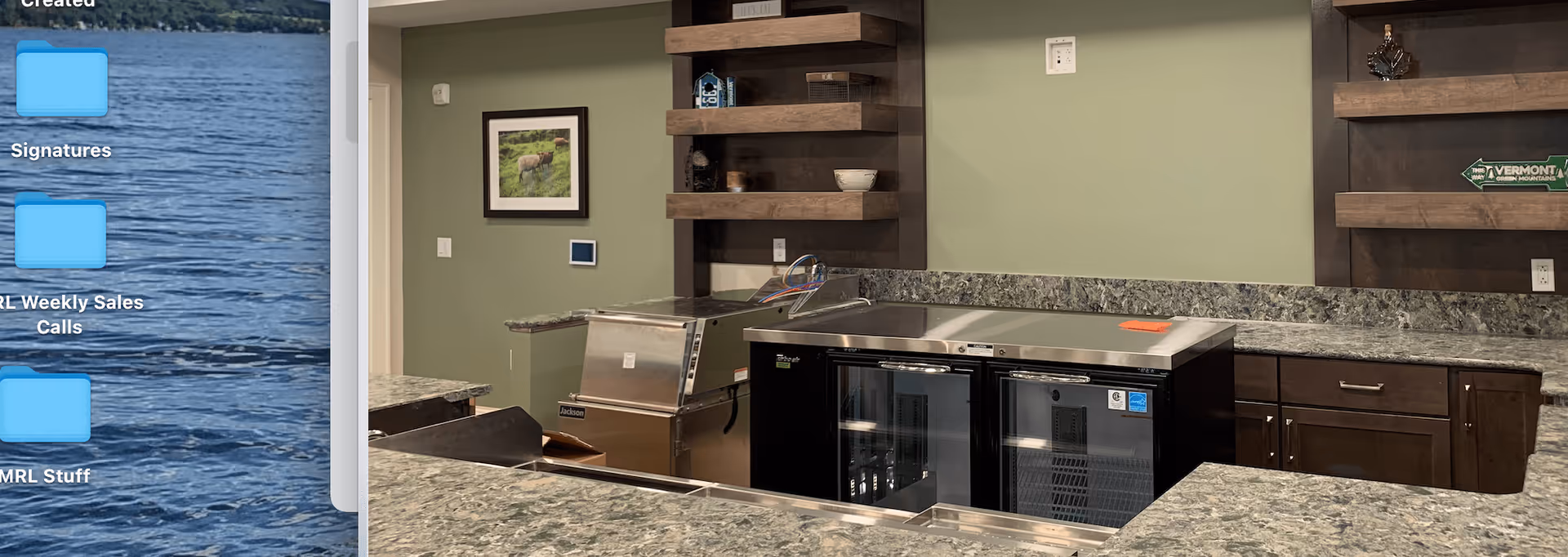 Interior view of a kitchen area with green walls, granite countertops, dark wooden cabinets, and open wooden shelves. There are two glass-door refrigerators under the counter and a stainless steel ice machine. A framed picture and decorative items are on the wall and shelves.