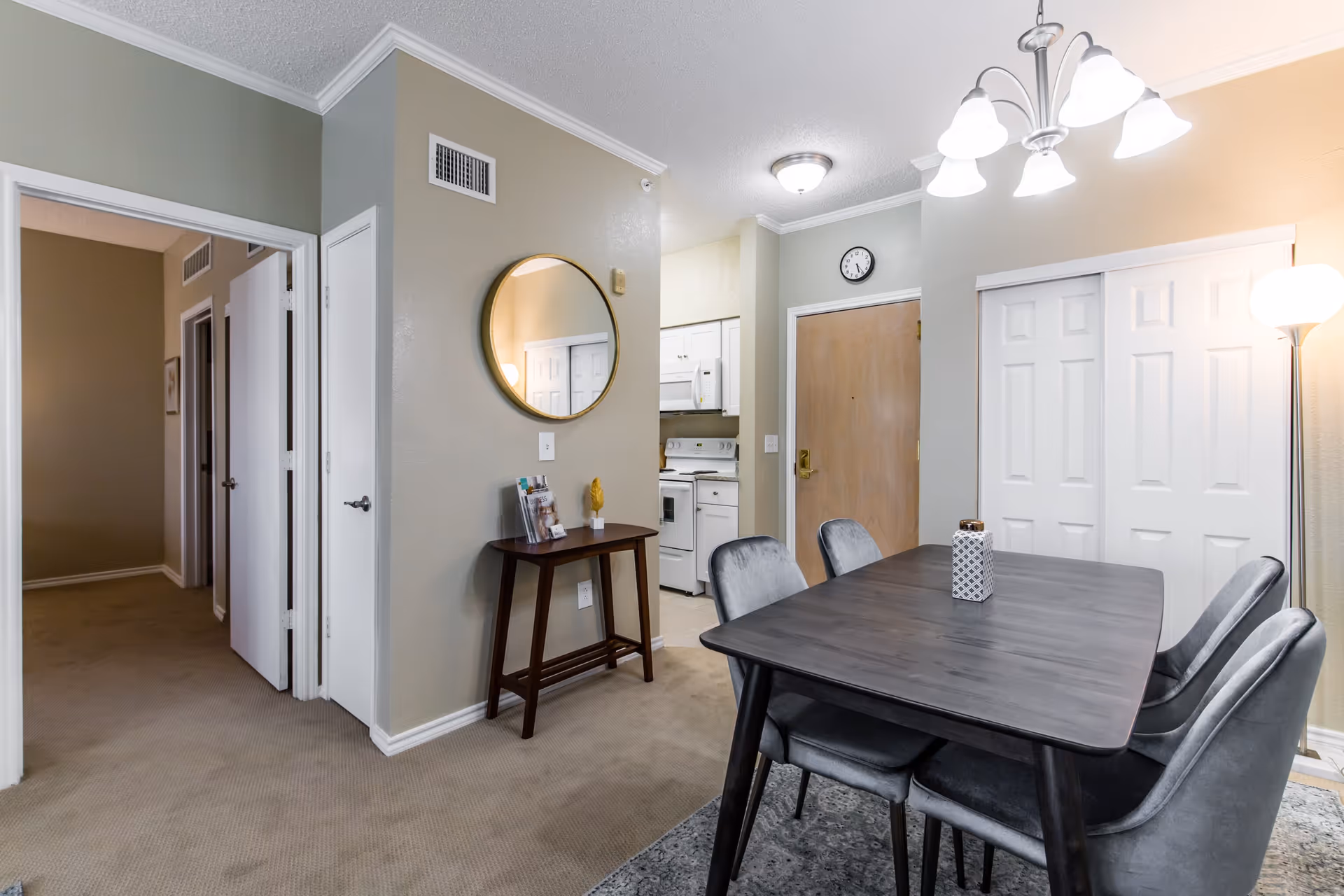 Dining area with a dark wood table and four chairs next to a small kitchen and entry door, with a round mirror on the wall.