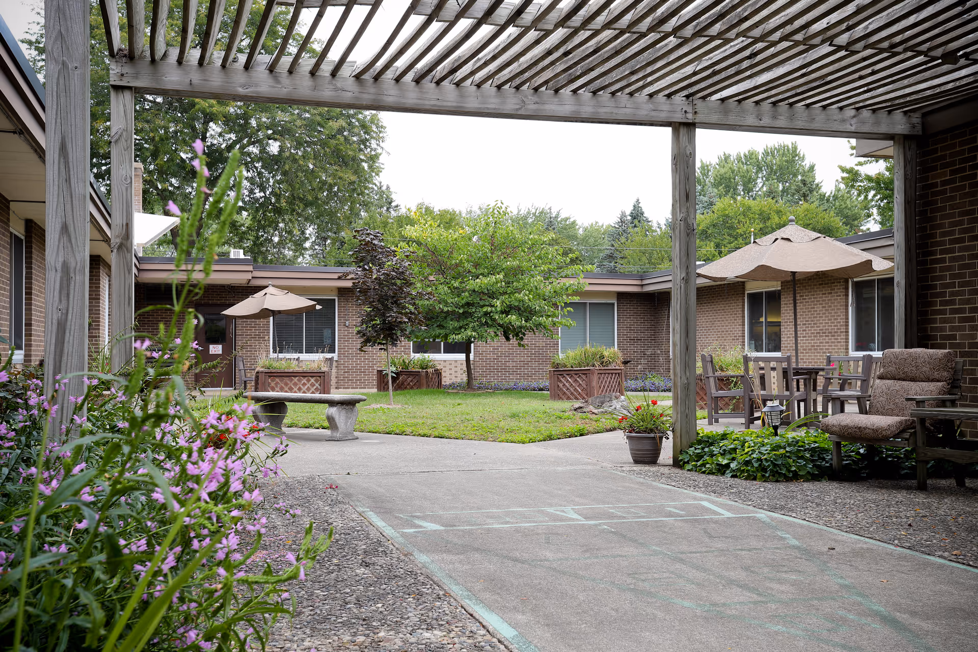 Outdoor courtyard area at Laurels of Hudsonville featuring a pergola overhead, concrete pathways, green grass, flower beds, potted plants, patio tables with umbrellas, and cushioned chairs surrounded by brick building walls and windows.