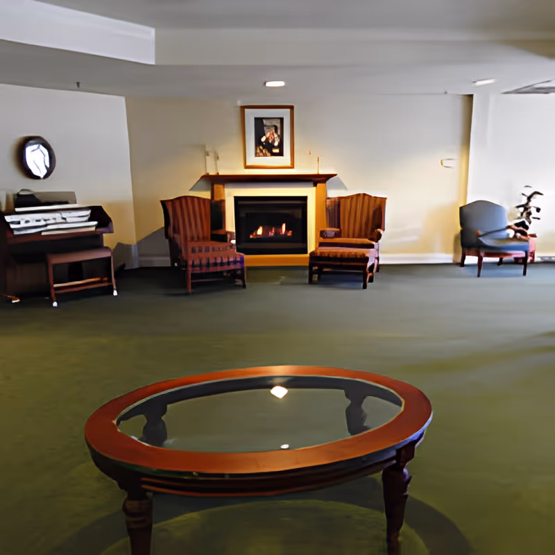 Sitting room with a round glass-top coffee table in the foreground and upholstered chairs arranged around a fireplace on a green carpet.