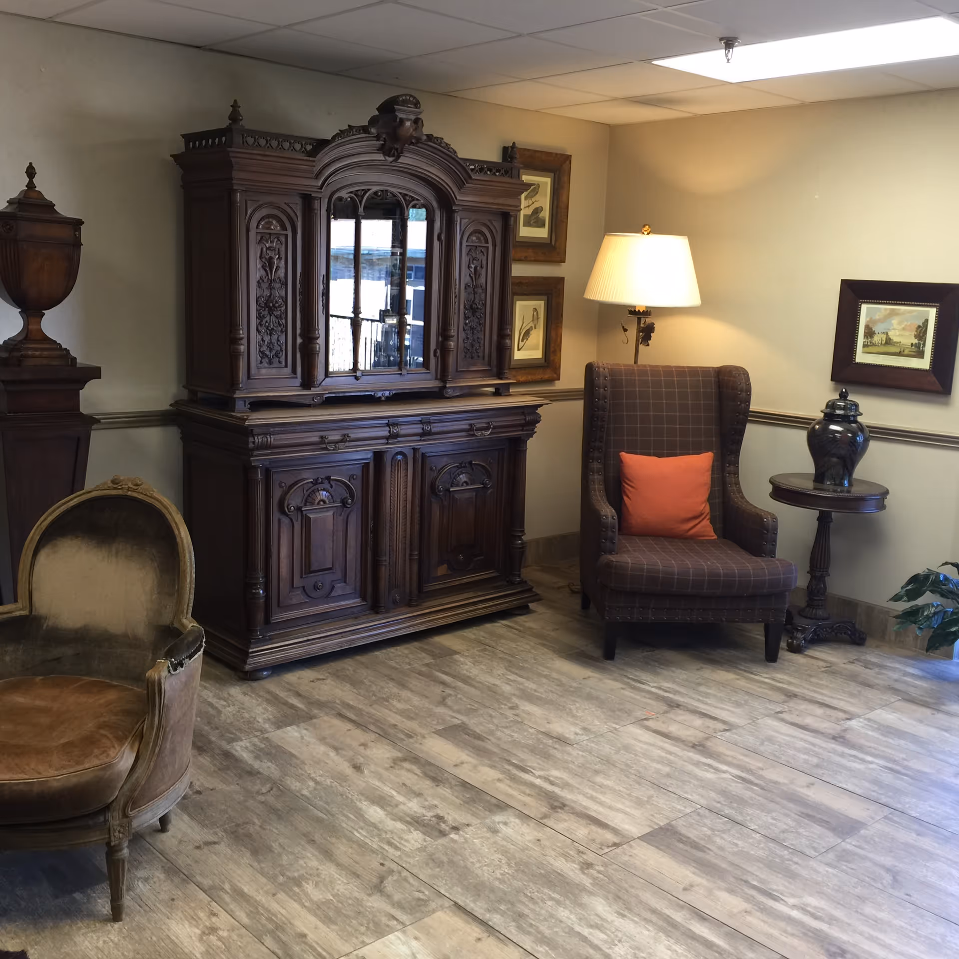 A cozy corner of a room featuring a vintage wooden cabinet with intricate carvings and mirrored panels, a brown upholstered armchair with an orange cushion, a small round wooden side table with a decorative vase, a floor lamp with a white shade, framed artwork on the walls, and a brown velvet armchair on the left side. The floor is covered with light wood-patterned tiles.