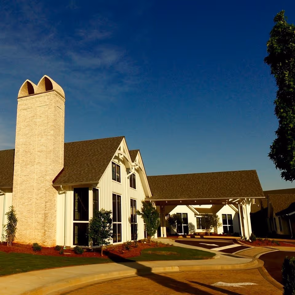 Front exterior of a white senior living building with a tall brick chimney and a covered entrance under a clear blue sky.