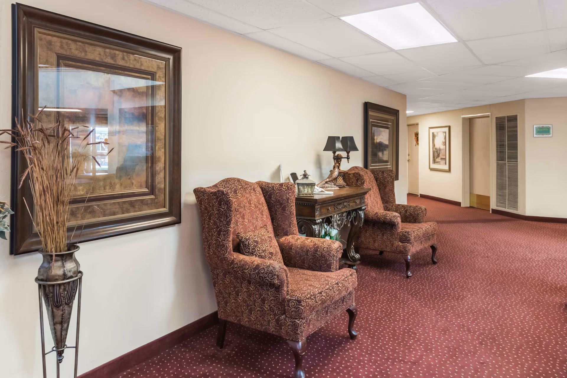 A hallway seating area in Asher Point Independent Living of Waco featuring two patterned upholstered armchairs with a decorative wooden side table between them. The table holds a lamp and some small decorative items. The walls are light-colored with framed artwork, and the floor is covered with a red carpet with a subtle dotted pattern. A tall vase with dried plants is placed near the wall on the left side.