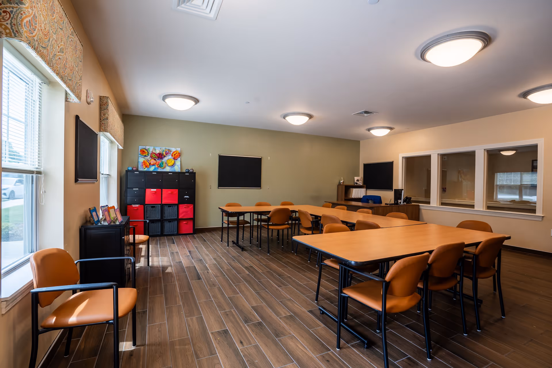 A well-lit room with several rectangular tables arranged in a U-shape, surrounded by orange chairs. The room has wood-look tile flooring, beige and green walls, and multiple ceiling lights. There are windows on the left side with patterned valances, a black storage unit with red and black bins against the far wall, and a small desk area with a computer and chair in the back right corner.