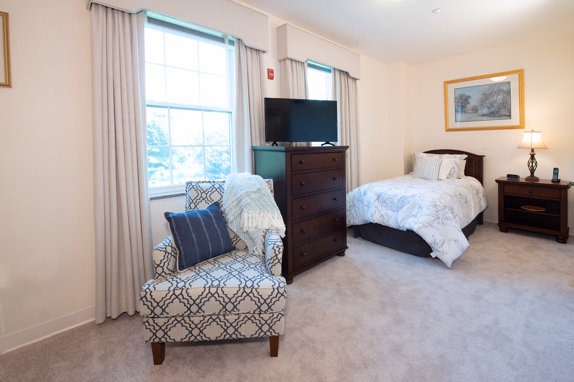 Bright furnished bedroom with a single bed, chest of drawers topped by a TV, patterned armchair by a window, and a nightstand with a lamp.