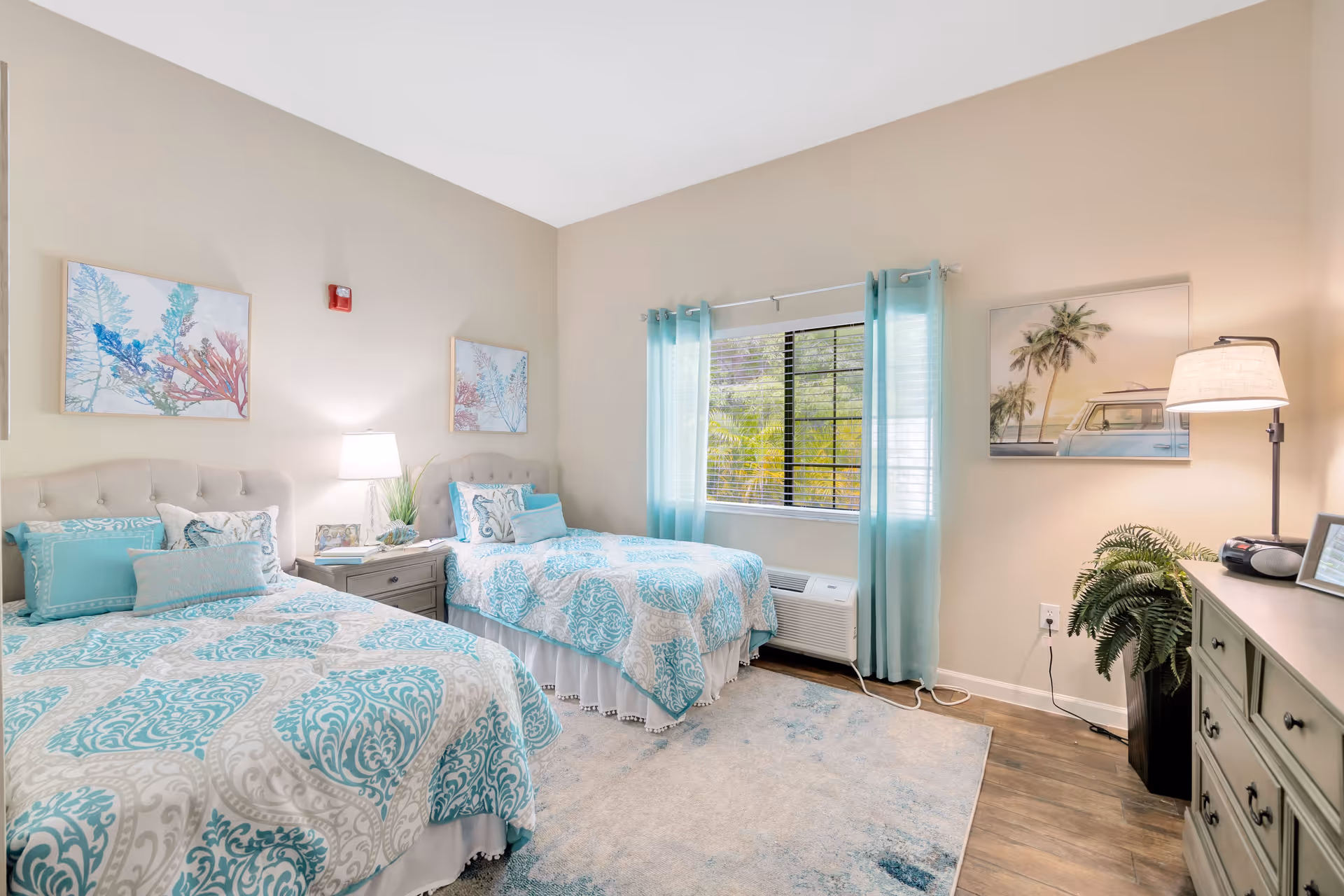 A bright and cozy bedroom with two beds featuring matching turquoise and white patterned bedding. The room has light beige walls adorned with framed artwork, a window with turquoise curtains, a nightstand with a lamp and decorative items, a dresser with a lamp and a plant, and wood flooring with a light area rug.
