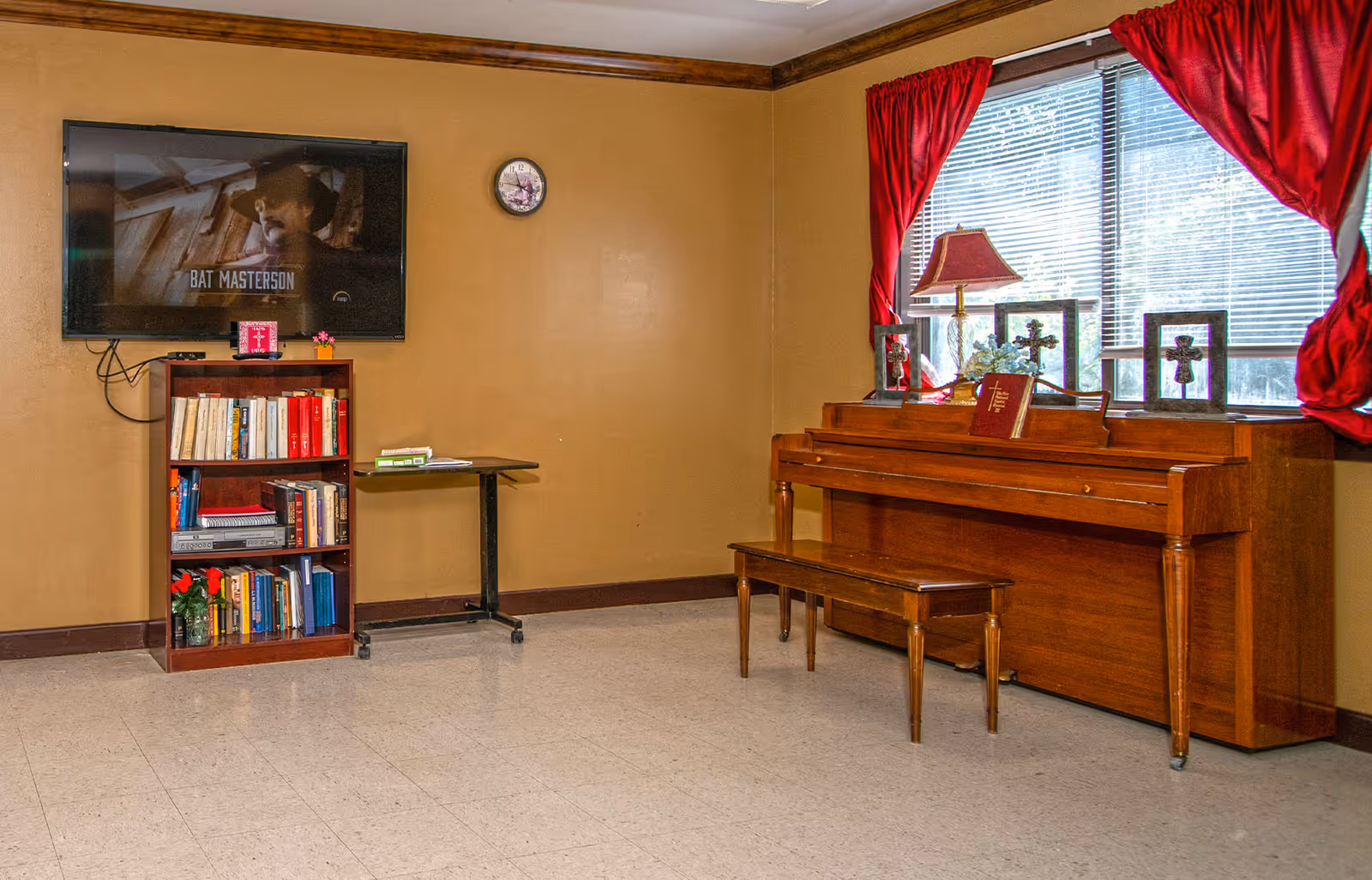 A room with a wooden piano and matching bench positioned under a window with red curtains. On top of the piano are three framed crosses, a lamp with a red shade, and a book. To the left, there is a small bookshelf filled with books and a flat-screen TV mounted on the wall above it. The walls are painted tan, and the floor is covered with light-colored tiles.