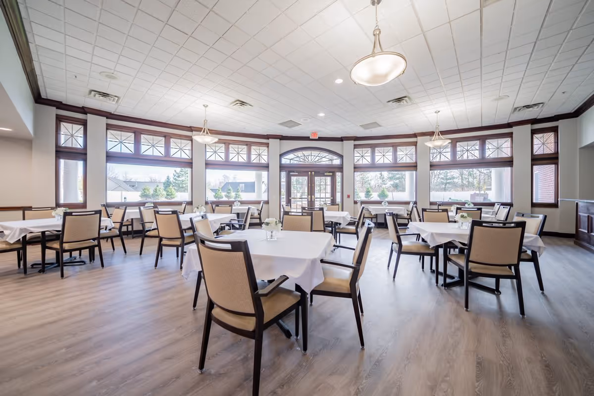 A spacious dining room with multiple tables covered in white tablecloths, each surrounded by beige cushioned chairs. The room has large windows allowing natural light to fill the space, and pendant lights hanging from a white tiled ceiling. The floor is a light wood laminate, and small floral centerpieces are placed on each table.