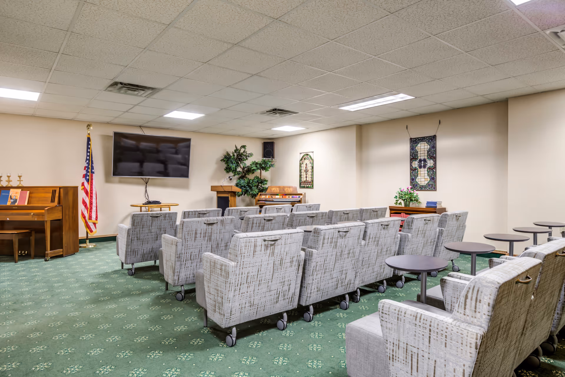A small auditorium or meeting room with rows of cushioned chairs on wheels facing a wall-mounted flat screen TV. There is a wooden piano and an American flag on the left side, a podium with a plant behind it, and decorative wall hangings. The room has a green carpet with a subtle pattern and a drop ceiling with fluorescent lights.