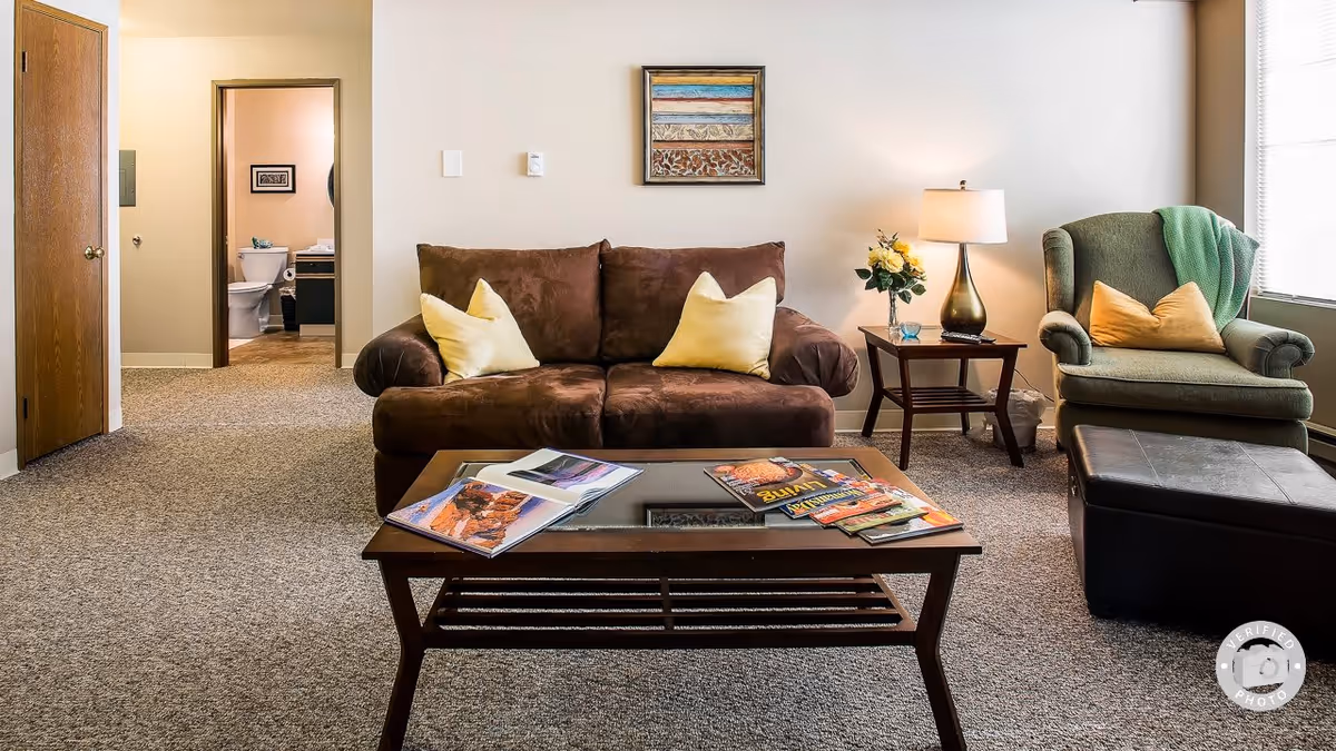 A cozy living room with a brown loveseat adorned with two yellow pillows, a green armchair with a yellow pillow and a green throw blanket, a wooden coffee table with magazines, and a side table with a lamp and a vase of flowers. In the background, there is a doorway leading to a bathroom with a visible toilet and sink.