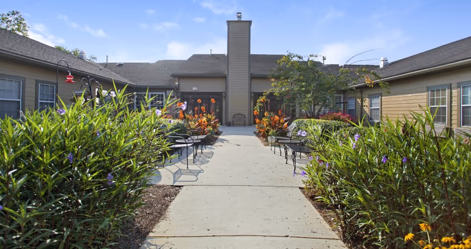 Outdoor courtyard area at American House Shallowford with a concrete walkway lined by green plants and flowers. There are black metal benches and chairs on both sides of the walkway. The building surrounds the courtyard with beige siding and multiple windows under a clear blue sky.