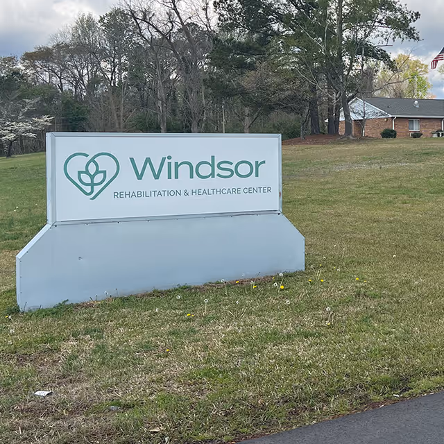 A freestanding sign on a grassy lawn that reads "Windsor Rehabilitation & Healthcare Center" with trees and part of the building behind it.