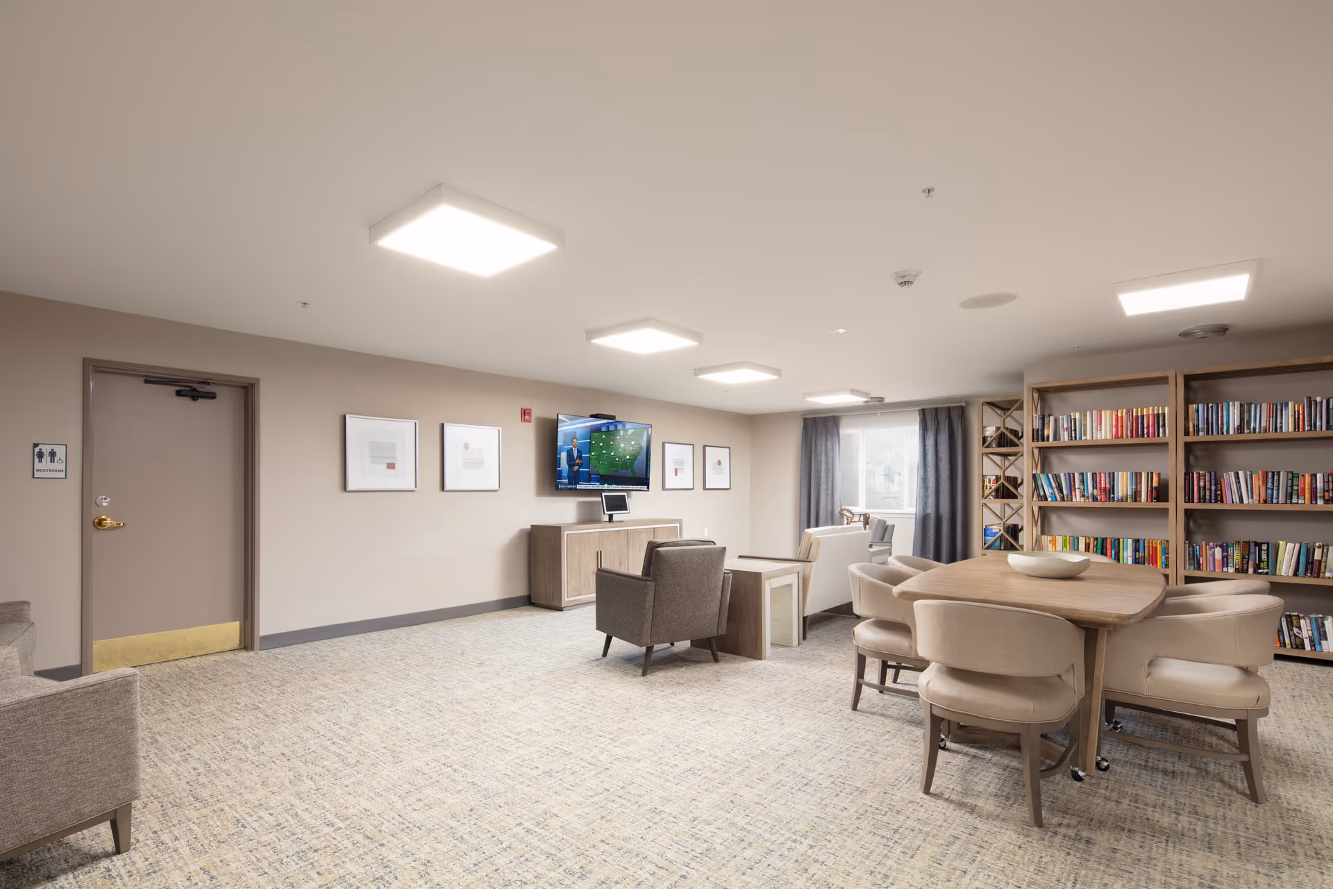 A spacious common area in a senior living facility with beige walls and carpeted floor. The room features a wooden table with six cushioned chairs, a bookshelf filled with books, two armchairs, a TV mounted on the wall showing a weather forecast, and framed pictures on the wall. There is a window with curtains letting in natural light and a door marked as a restroom.