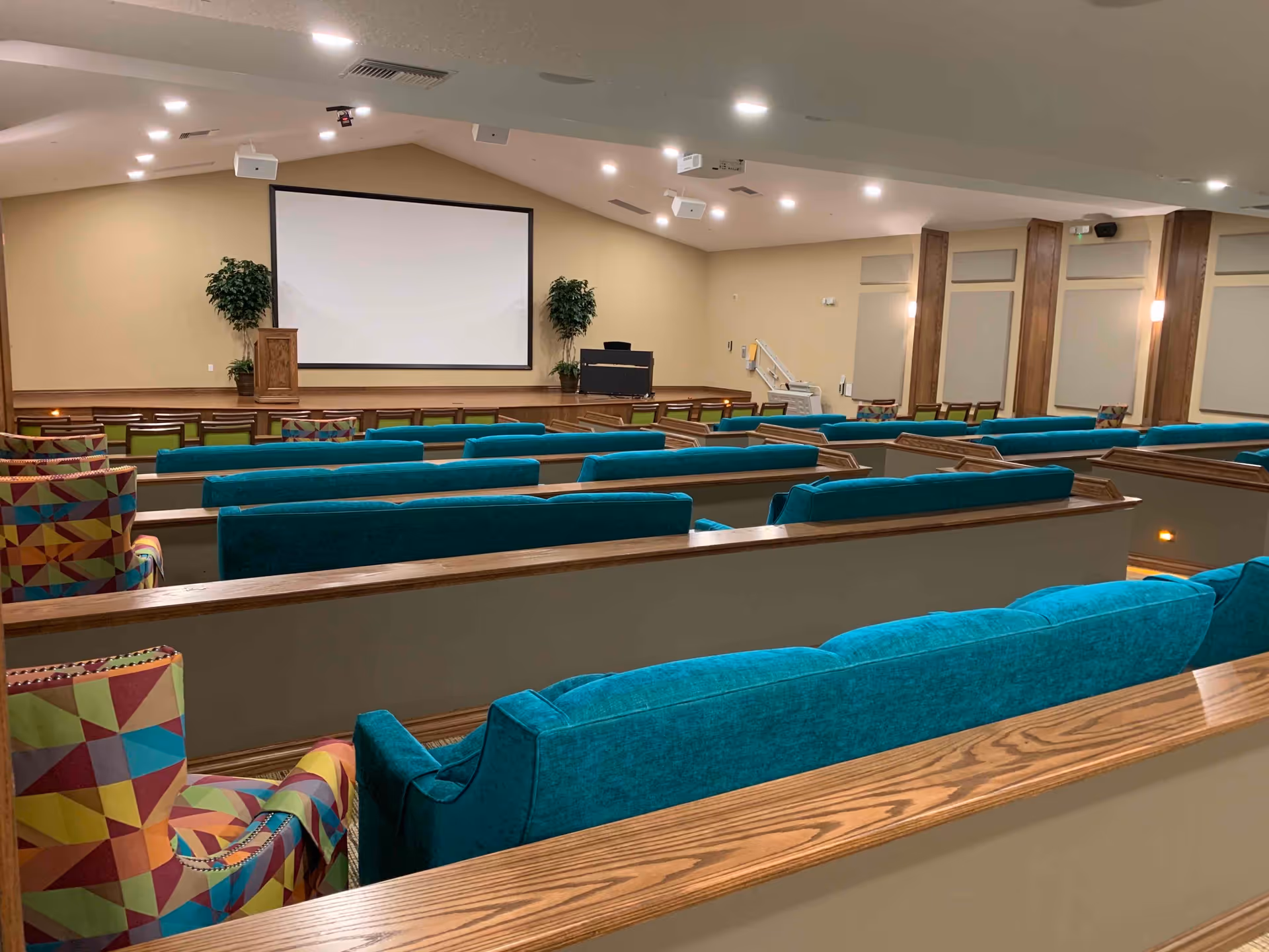 Auditorium-style room with rows of teal upholstered seats and patterned chairs facing a stage with a large projection screen.