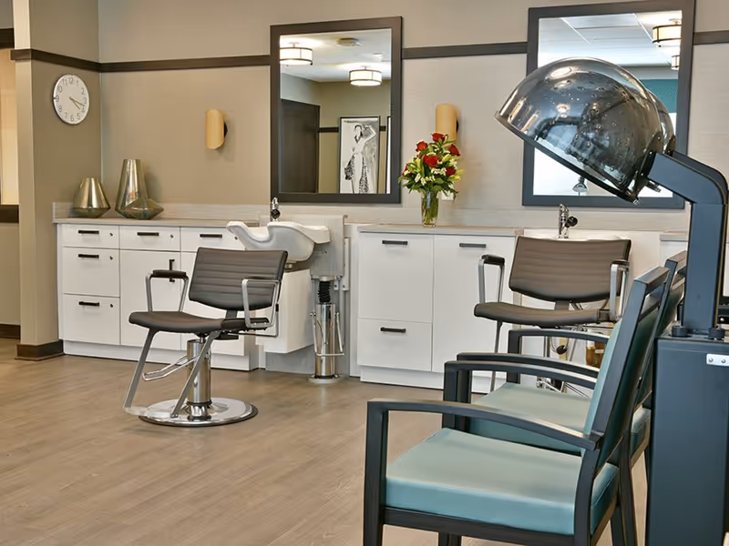 Interior of a senior living facility hair salon with two salon chairs in front of mirrors, a hair washing station, a hair dryer, and a couple of waiting chairs. There are decorative vases and a bouquet of flowers on the counter, and a clock on the wall.