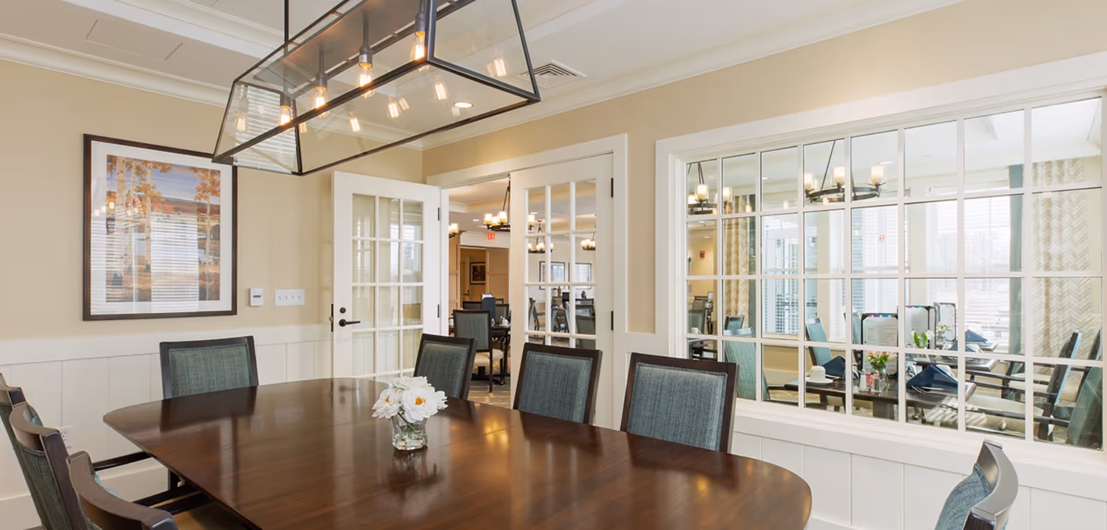 Bright dining room with a long wooden table, upholstered chairs, a central light fixture, and a view into an adjacent room through interior windows.