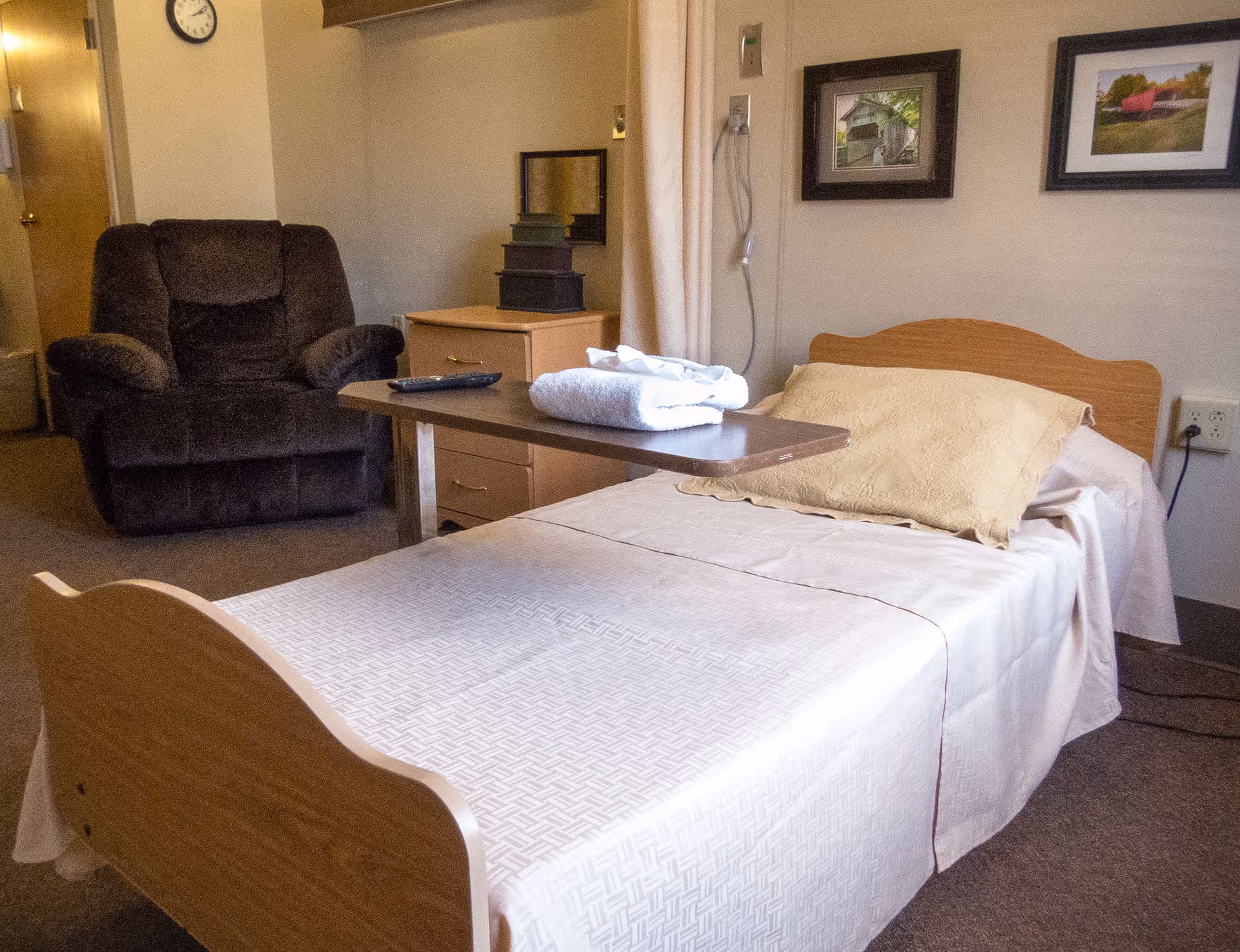 A small bedroom in a senior living facility featuring a single bed with a beige pillow and white bedspread, a brown reclining chair, a wooden nightstand with stacked boxes, a wall clock, two framed pictures on the wall, and a movable overbed table with folded towels and a remote control on it.